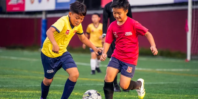 Two children in JSSL kit chasing a football on an artificial pitch