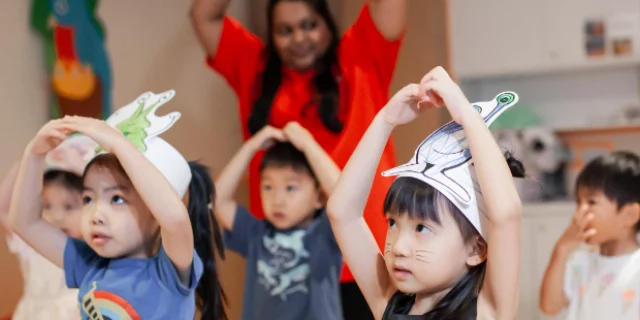 Young children in paper animal hats copying a teacher’s arm movements in a drama class