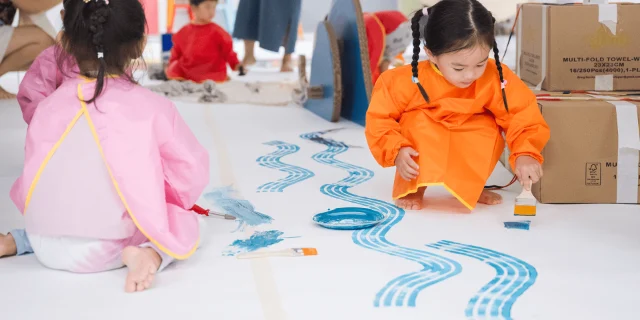 Two young children in painting smocks printing blue patterns on paper on the floor