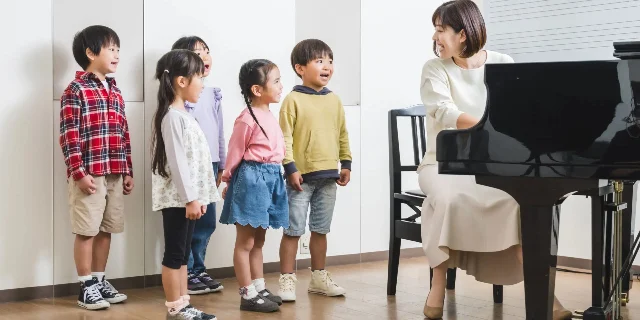 Five young children standing by a grand piano while a teacher plays